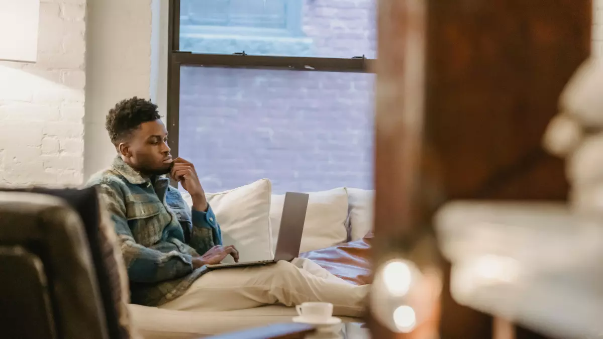 A man is sitting on a couch in a living room, looking at a laptop.