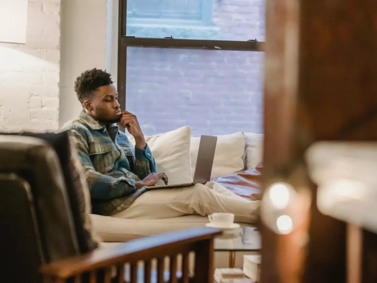 A man is sitting on a couch in a living room, looking at a laptop.