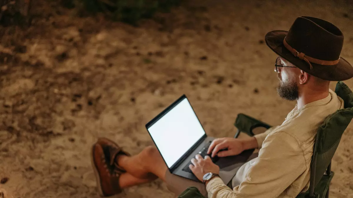 A man is sitting on a chair, working on a laptop, in an outdoor setting. He is wearing a hat and is looking at the screen.