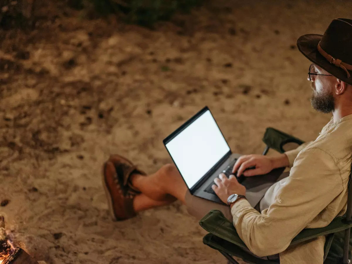 A man is sitting on a chair, working on a laptop, in an outdoor setting. He is wearing a hat and is looking at the screen.