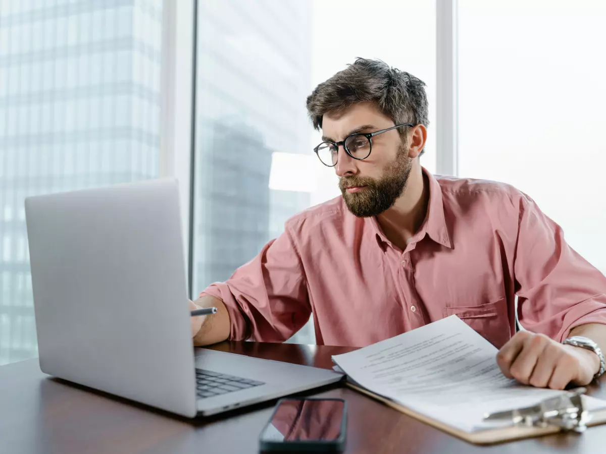 A man with glasses, a beard and a pink shirt is sitting at a desk in front of a laptop and working on a laptop. He is serious and focused. He is likely a trader. There is a newspaper on the desk in front of him.