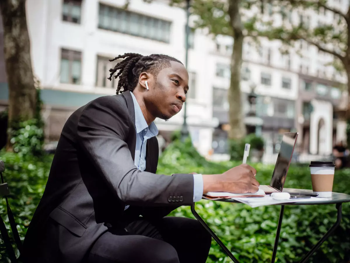 A young man in a suit, with dreadlocks, is sitting on a park bench. He is working on a laptop, taking notes on a pad, and has a cup of coffee on the table next to him. There are trees and buildings in the background. 