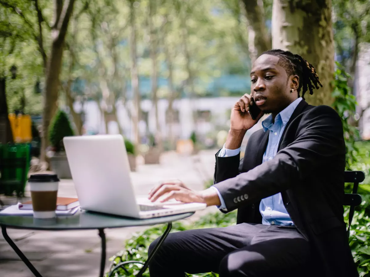 A man in a suit is sitting at a table in a park, working on his laptop and talking on his phone.