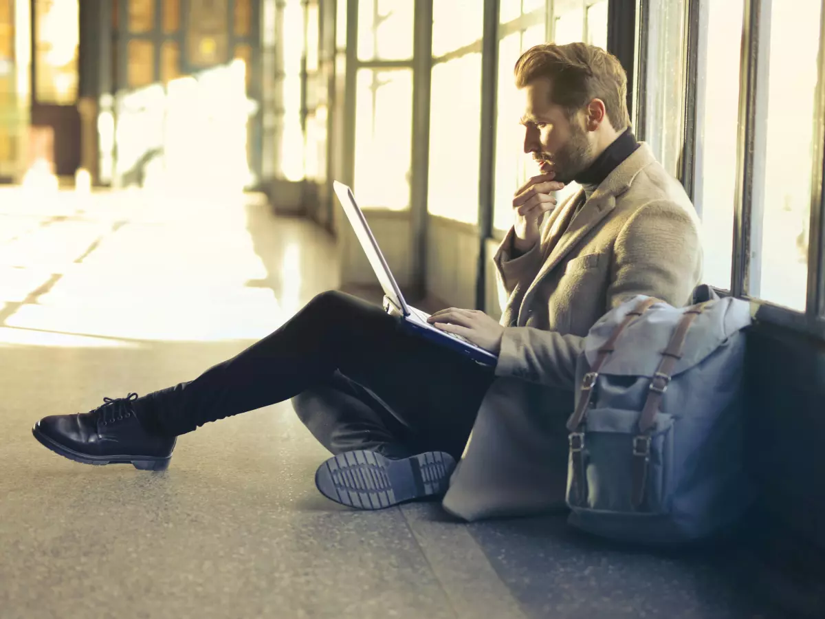 A man with a beard, wearing a blazer and black pants, is sitting on the floor in an indoor setting, focused on his laptop, which is resting on his legs.  He has a backpack next to him. The room is illuminated by a window in the background. 