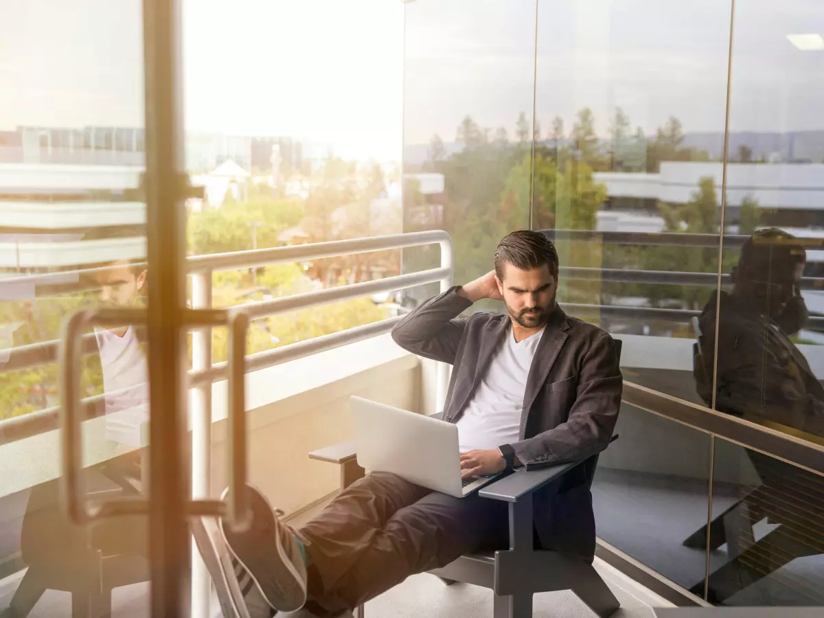 A man in a suit sits on a balcony, looking stressed as he works on his laptop.