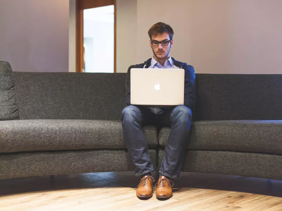 A man in a casual outfit is sitting on a couch, working on a laptop.