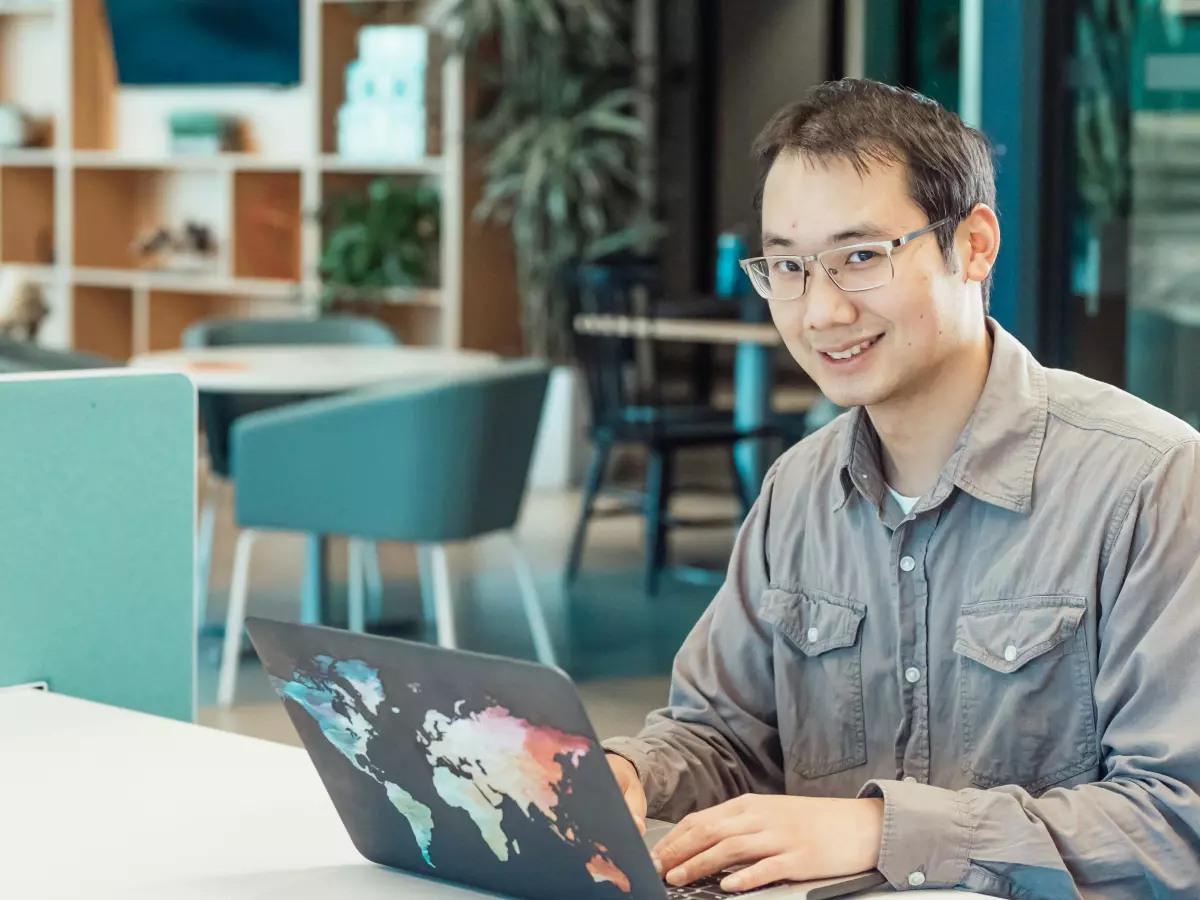 A young man wearing a grey button-up shirt with glasses is smiling as he uses a laptop in an office setting.  The laptop is open and a digital world map is displayed on the screen.