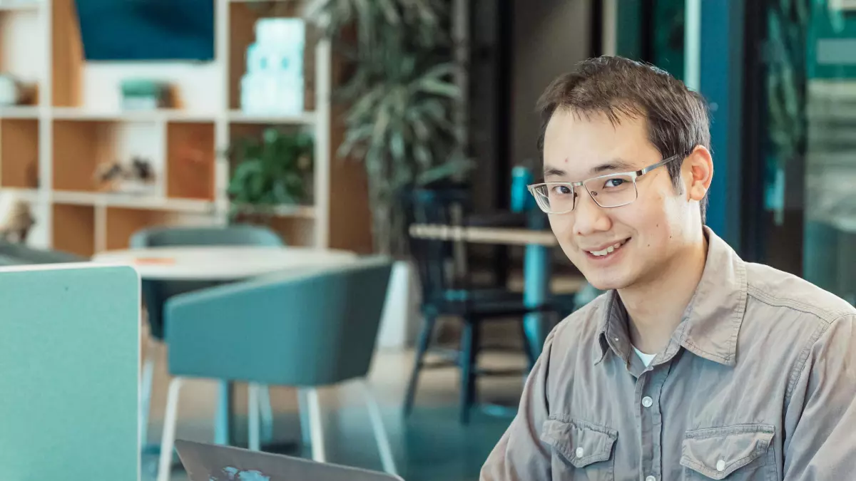 A young man wearing a grey button-up shirt with glasses is smiling as he uses a laptop in an office setting.  The laptop is open and a digital world map is displayed on the screen.