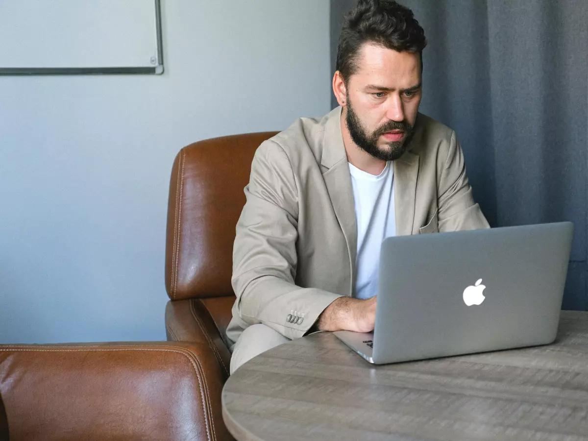 A man in a beige jacket is working on his laptop in a living room. He has a beard and is looking at the laptop.