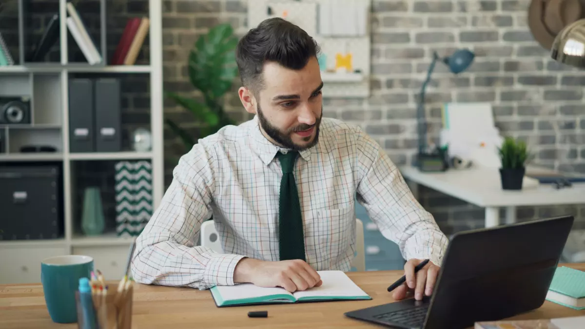 A man in a white shirt and tie is working on a laptop in a modern office. He is looking at the screen with a focused expression.