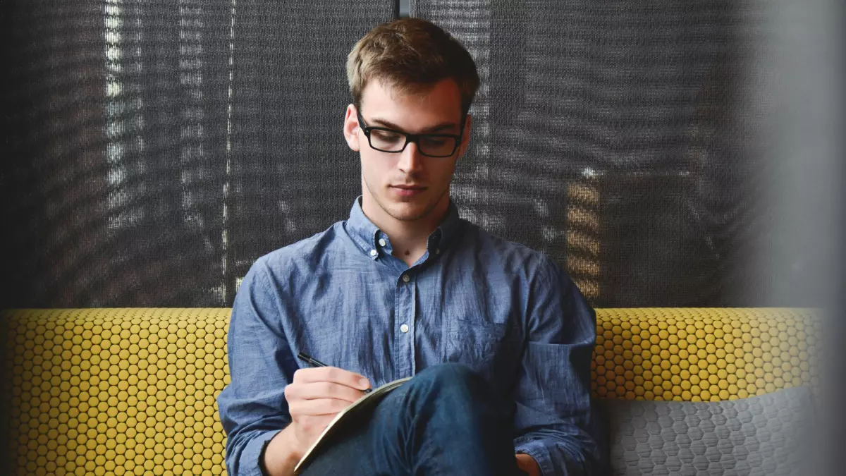 A young man in a blue shirt and jeans is sitting on a yellow couch, writing in a notebook. He is wearing glasses and has a serious expression on his face.