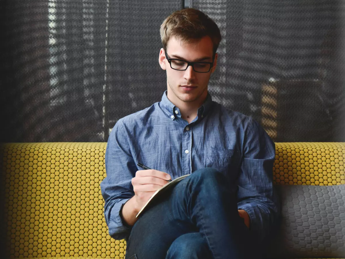 A young man in a blue shirt and jeans is sitting on a yellow couch, writing in a notebook. He is wearing glasses and has a serious expression on his face.