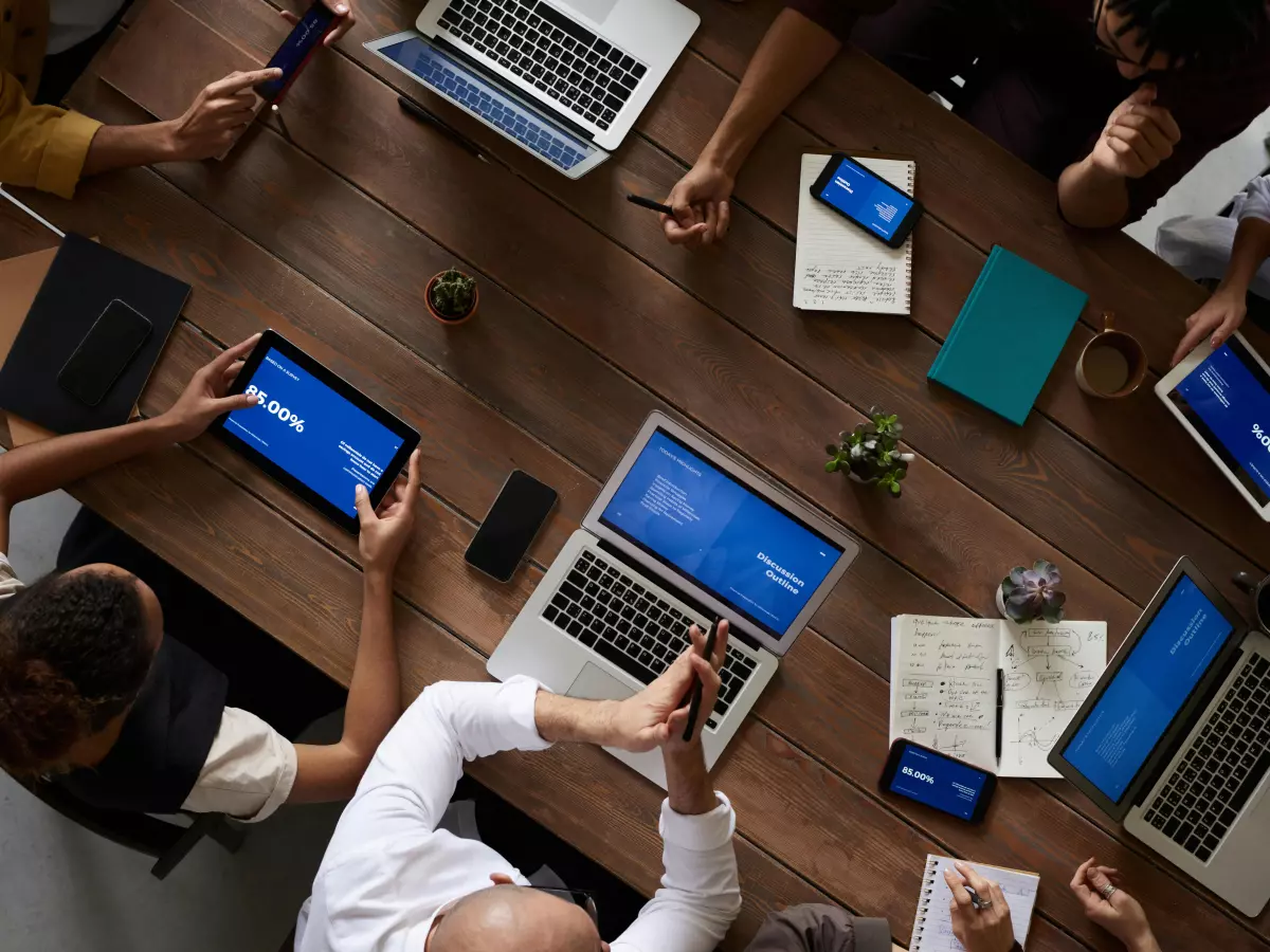 A diverse group of people are sitting around a table, working on their laptops and tablets. The scene is dynamic and engaging, showcasing the use of technology in a business setting.