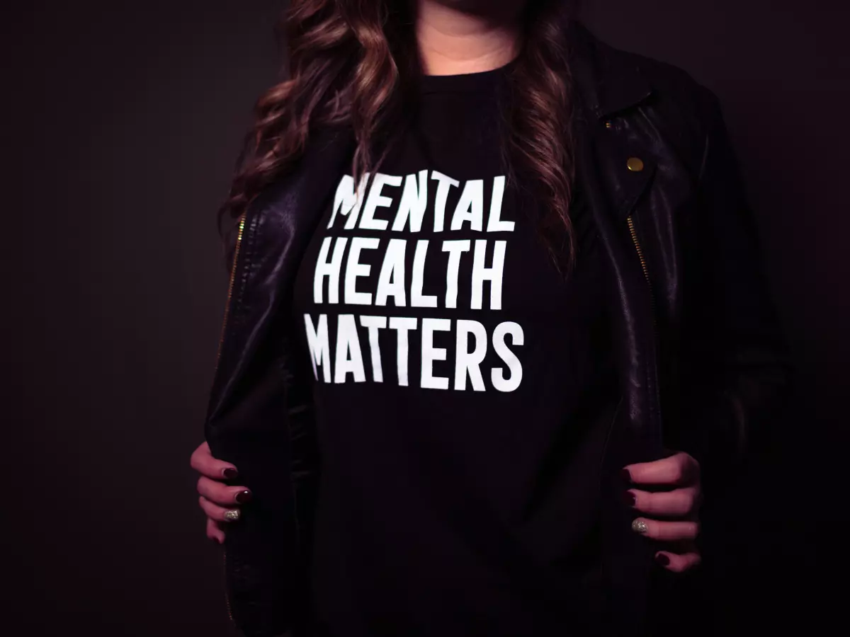 A woman wearing a black leather jacket and a black t-shirt with the words 'Mental Health Matters' printed on it. She is standing in front of a dark background.