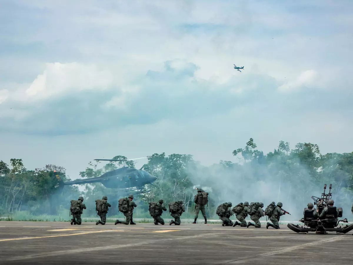 A group of soldiers in camouflage uniforms are standing in a line on a runway. There are two helicopters in the background, one of which is taking off. There is smoke in the air.