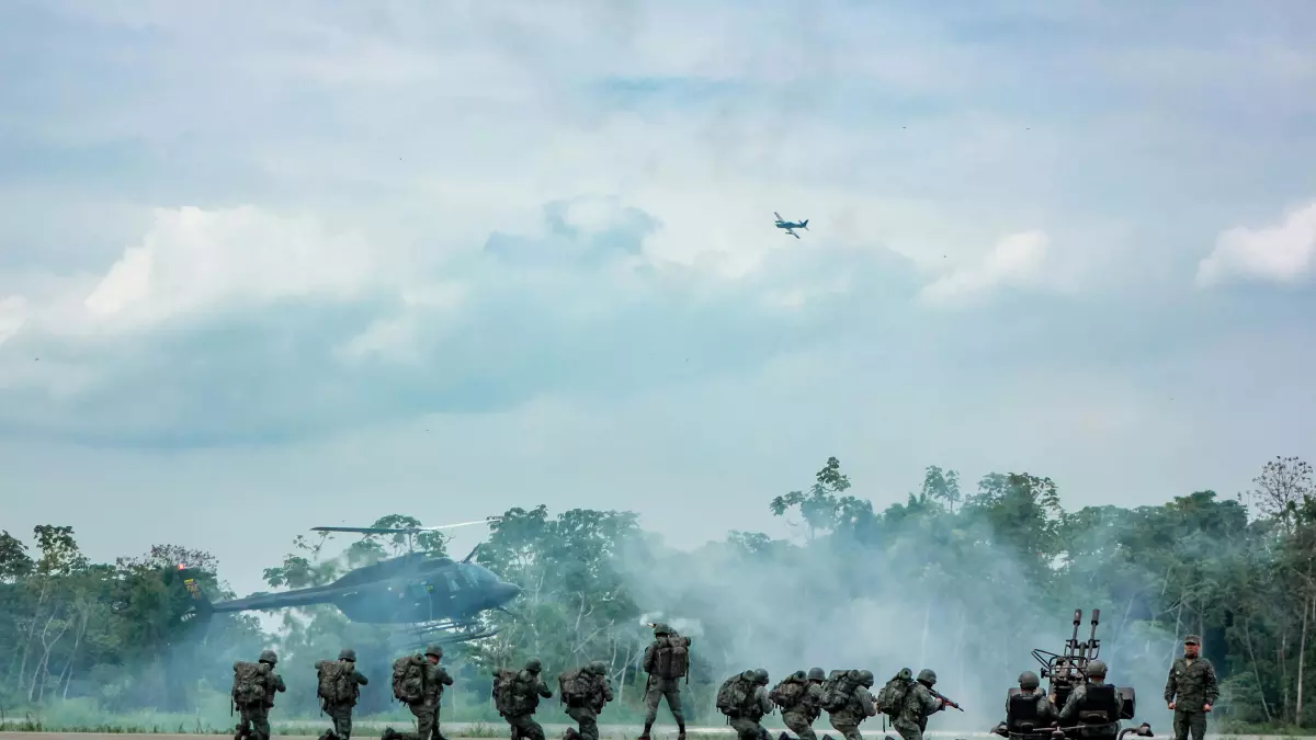 A group of soldiers in camouflage uniforms are standing in a line on a runway. There are two helicopters in the background, one of which is taking off. There is smoke in the air.