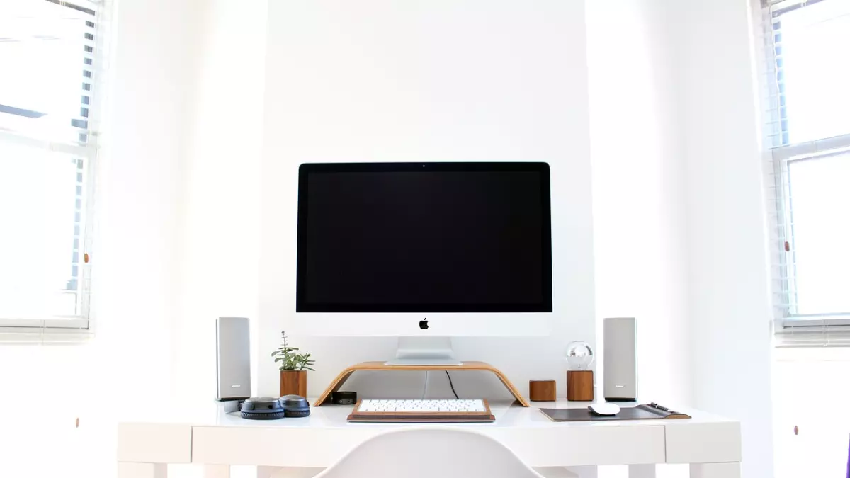 A white desk with a white iMac, a keyboard, a mouse, and two speakers. There is a plant in a pot on the desk, and a window behind the computer.