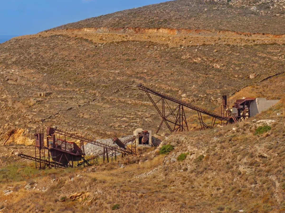 A wide shot of a quarry with rusty mining equipment and a conveyor belt, set against a dry, hilly landscape.