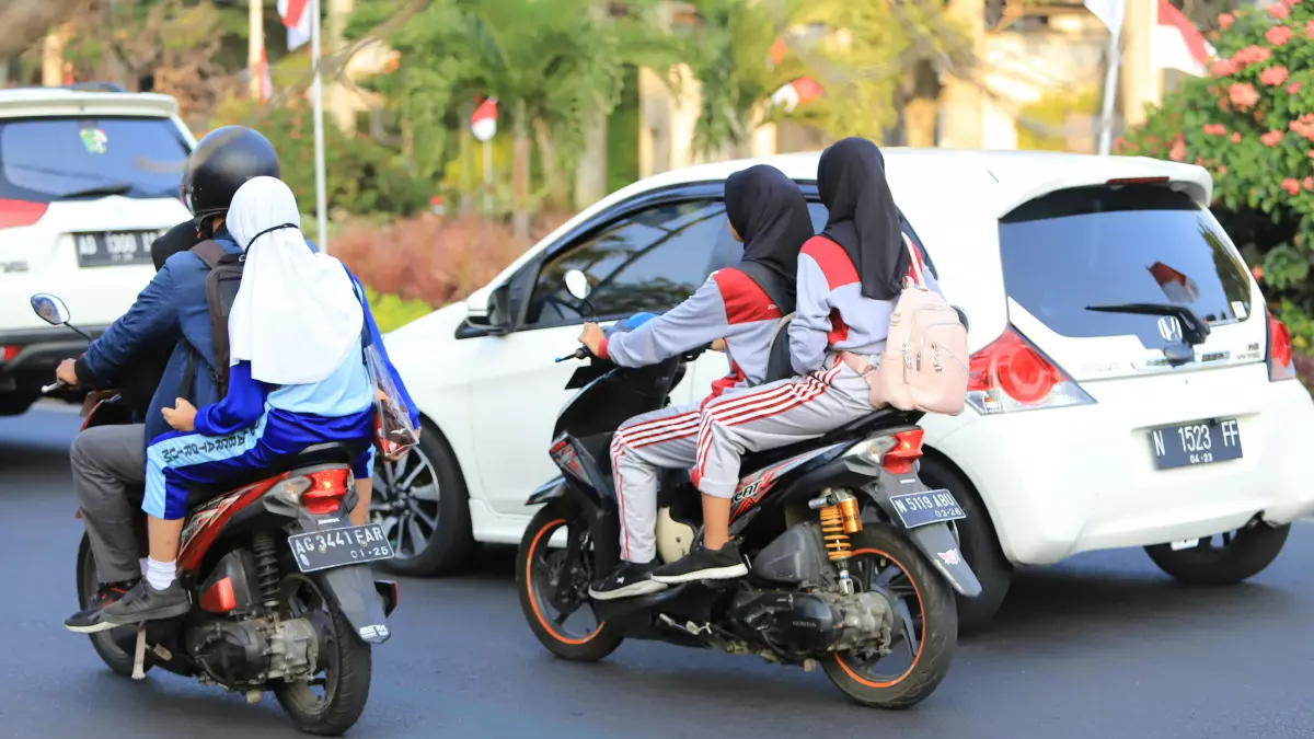 A scooter with two people on it, a woman and a girl, both wearing helmets, are driving on the road in a busy street.