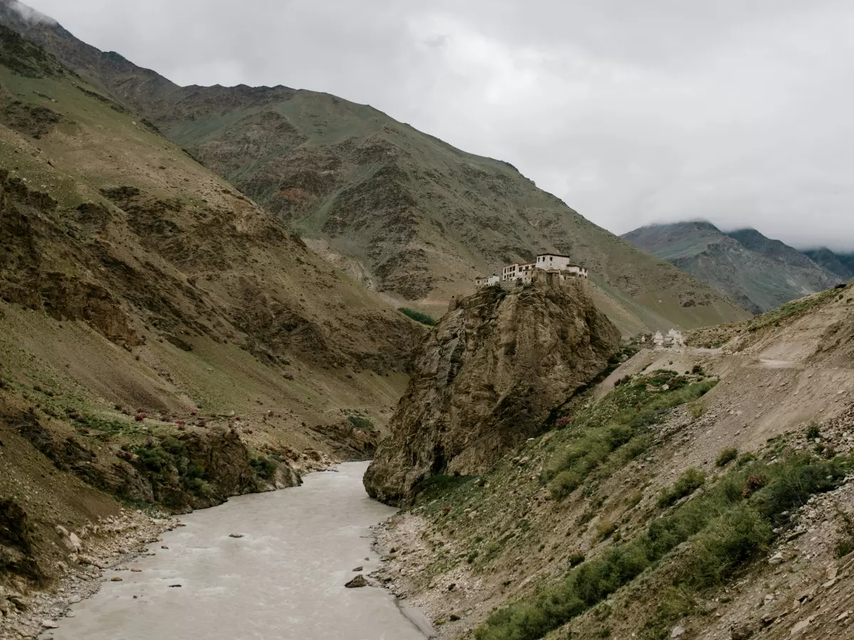 A photo of a winding road through a mountainous region, with a large rock in the foreground and a steep mountain range in the background. The road is paved and appears to be in good condition, but the surrounding landscape is rugged and barren.