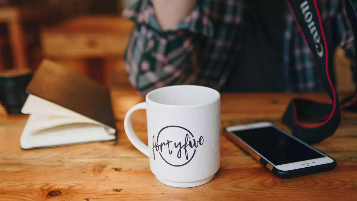 A white mug with a black logo on it sits on a wooden table next to a smartphone. A person