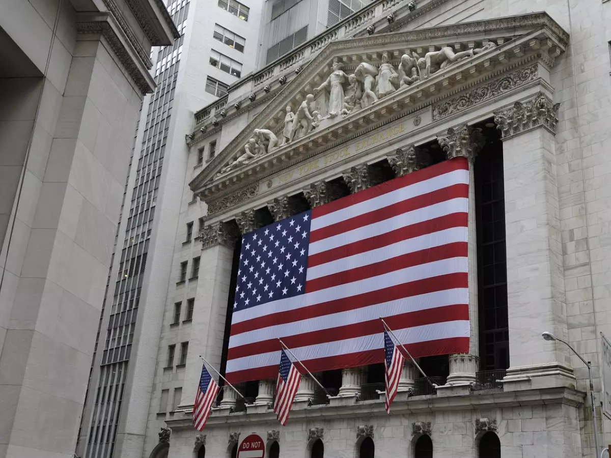 A low-angle shot of the facade of the New York Stock Exchange building with an American flag draped across it. The flag is seen prominently against the backdrop of a grey marble building.