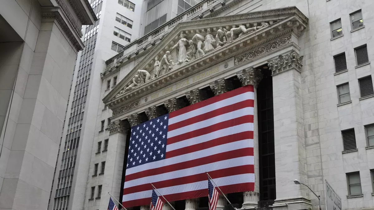 A low-angle shot of the facade of the New York Stock Exchange building with an American flag draped across it. The flag is seen prominently against the backdrop of a grey marble building.