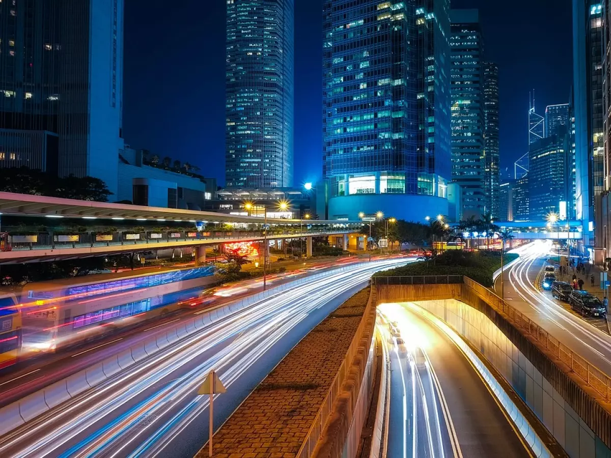 A night shot of a city street with cars driving through a tunnel with light trails. The shot is taken from above and shows the tall skyscrapers in the background.