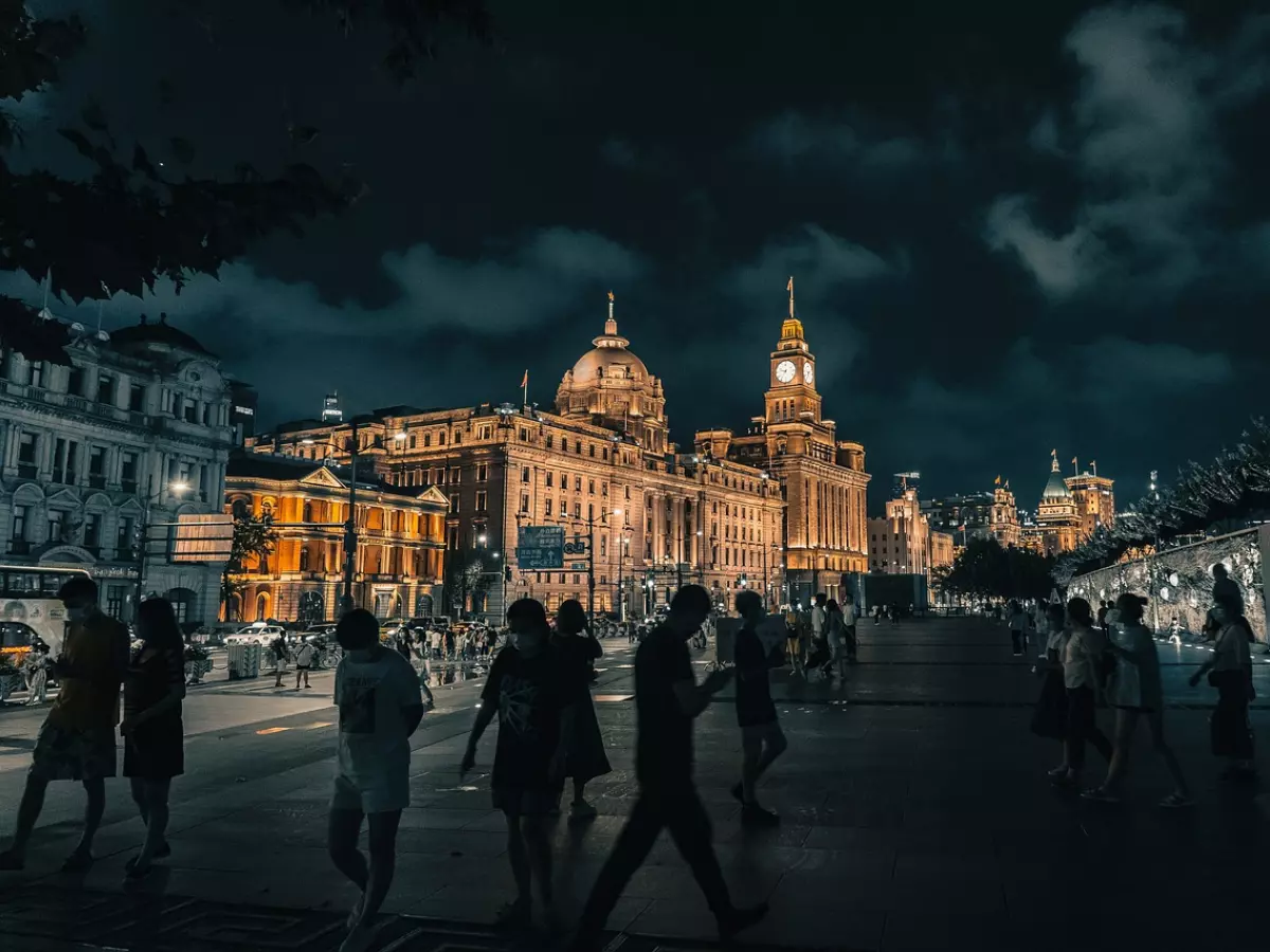 A night shot of a crowded street in Shanghai, with people walking past a grand building illuminated in warm light. The cityscape is a mix of modern and historical architecture, creating a sense of dynamism and cultural richness.