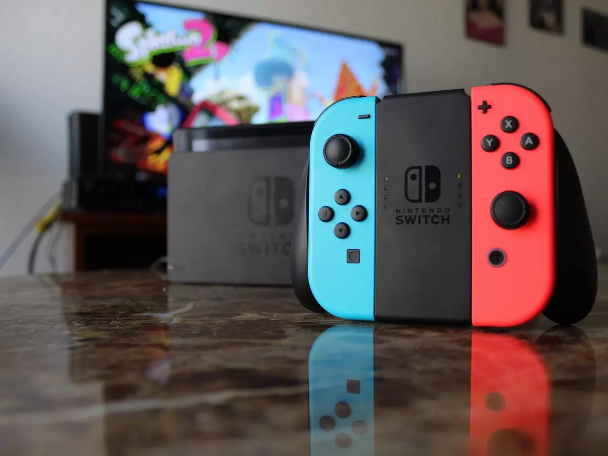 A Nintendo Switch console with a Joy-Con controller in the foreground, a TV screen in the background, and a marble table surface.