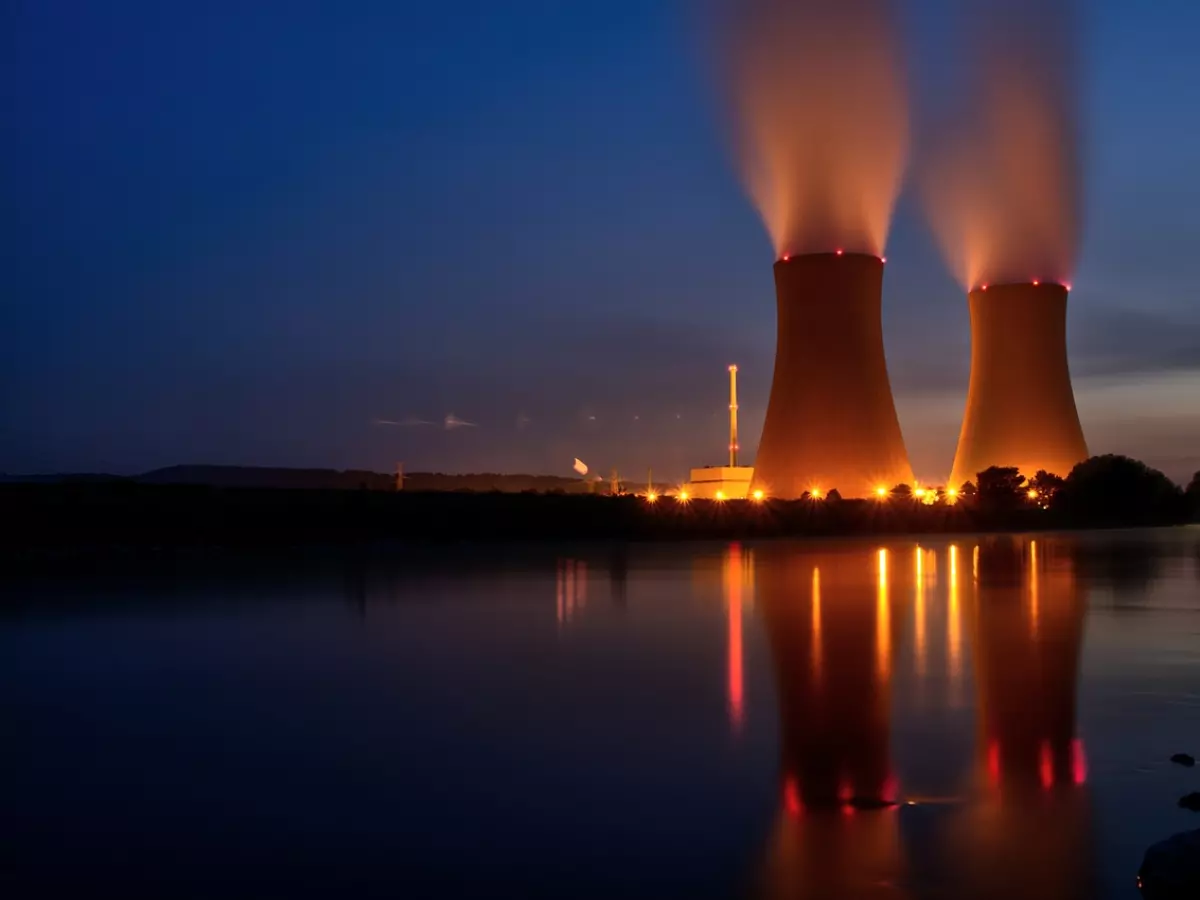 Two tall cooling towers of a nuclear power plant stand illuminated against a dark sky. The towers are reflecting in the still water, with faint clouds in the background. The image has a dramatic and almost ominous feel, hinting at the power and potential danger of nuclear energy.