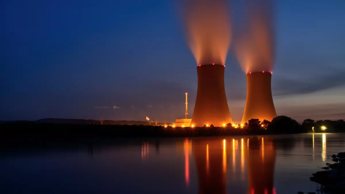 Two tall cooling towers of a nuclear power plant stand illuminated against a dark sky. The towers are reflecting in the still water, with faint clouds in the background. The image has a dramatic and almost ominous feel, hinting at the power and potential danger of nuclear energy.