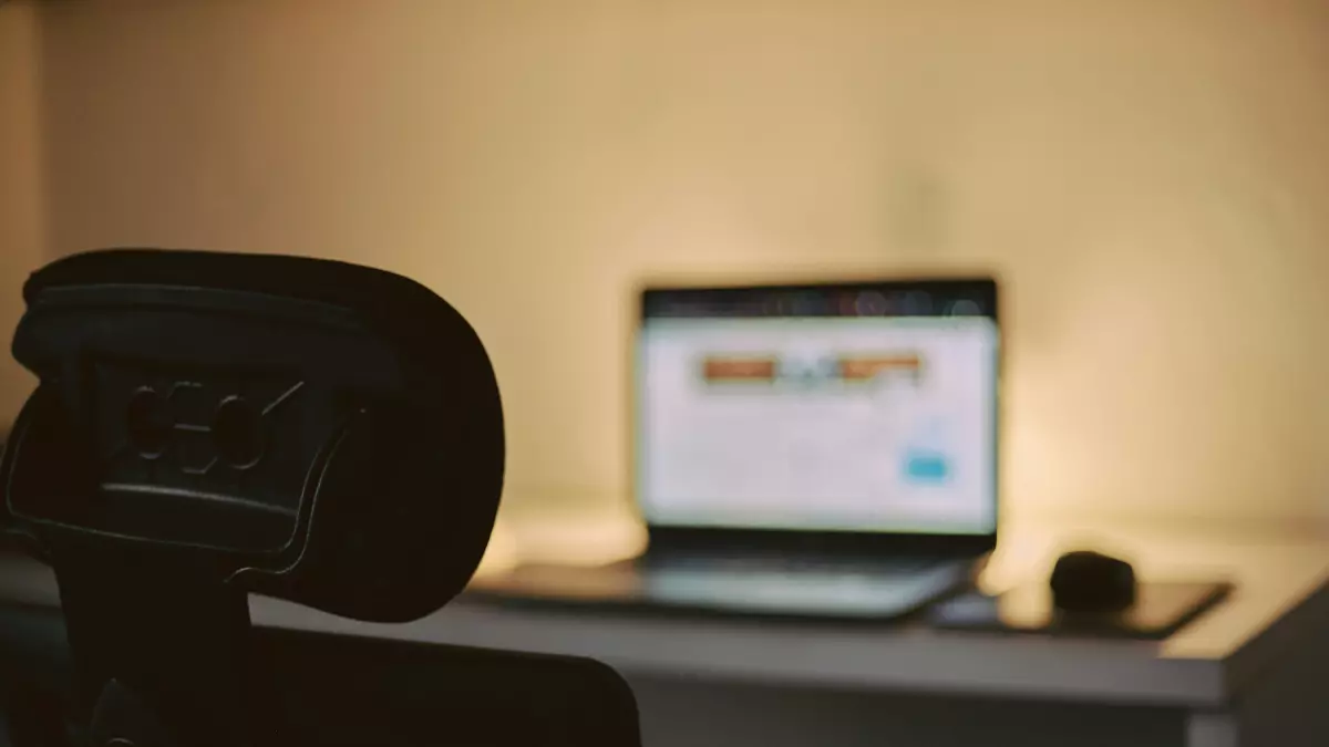 A laptop is on a desk in a dimly lit room, with the back of a chair in the foreground.