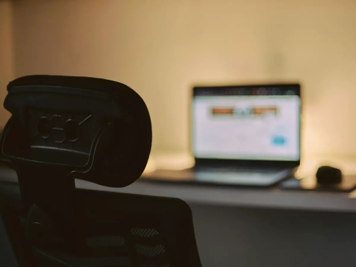 A laptop is on a desk in a dimly lit room, with the back of a chair in the foreground.