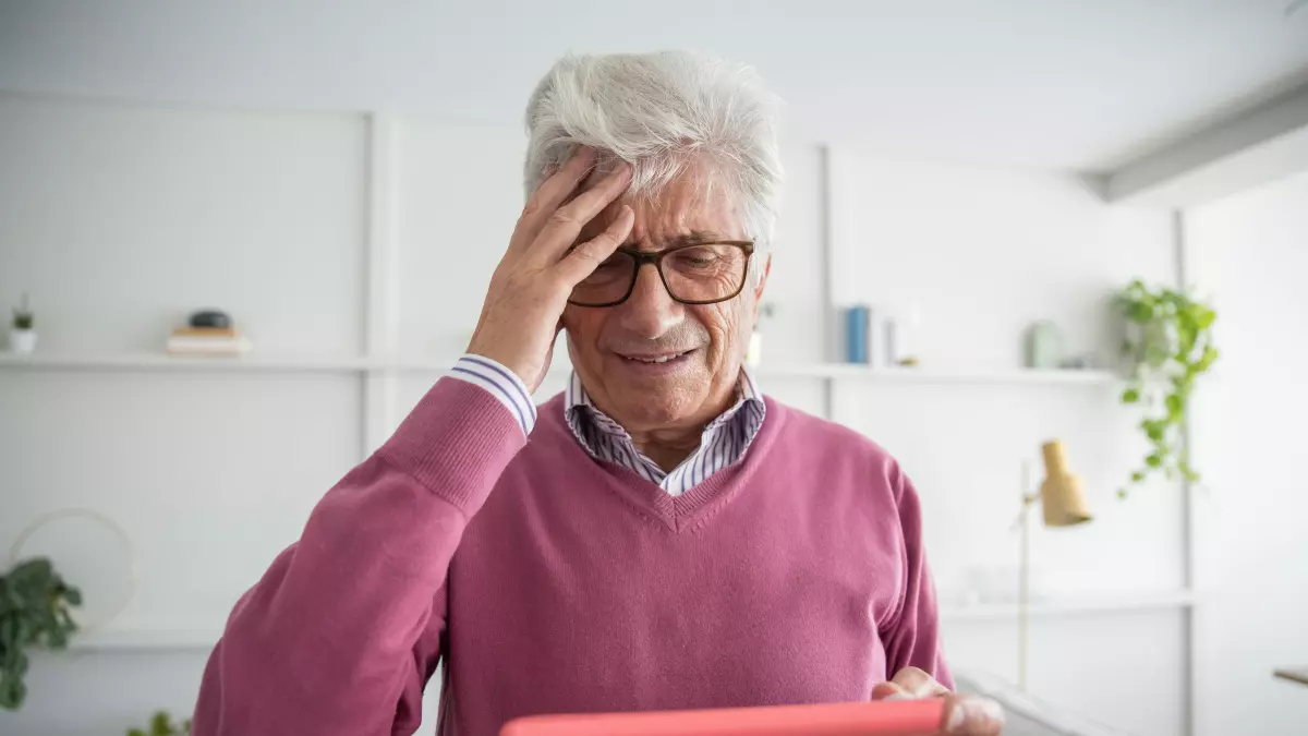 An elderly man with a distressed expression holds a tablet in his hand, while putting his hand on his forehead. 