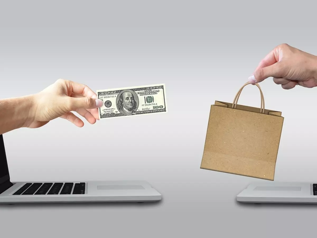 Two hands are reaching out of laptop screens. One hand is holding a $100 bill and the other hand is holding a brown paper bag.