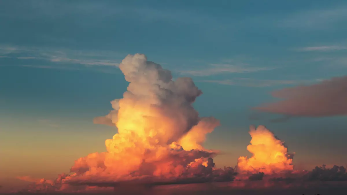 A dramatic sky with a large, puffy cumulus cloud illuminated by the setting sun, casting a warm orange glow. The cloud appears to be growing and expanding, with a smaller cloud to the right. The sky is a gradient of blue and orange, with a hint of pink.