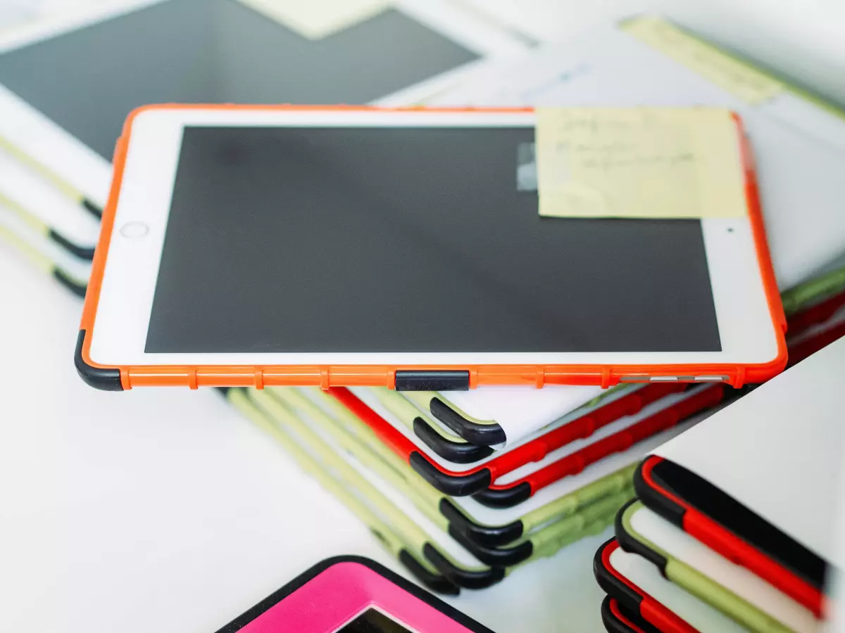 A close-up shot of an iPad tablet, showcasing its sleek design and vibrant orange case. The tablet is resting on a stack of other tablets, suggesting a modern learning environment.