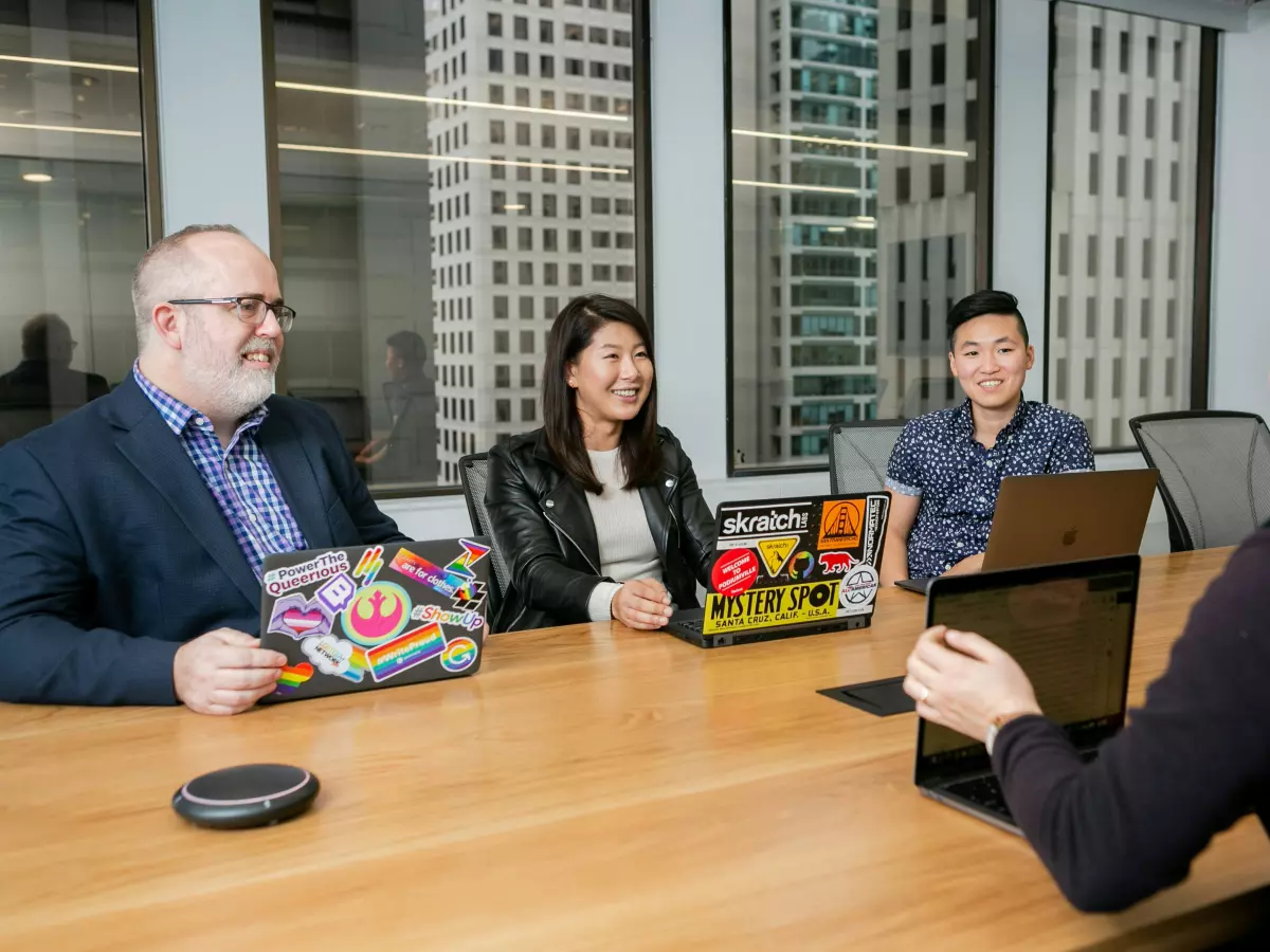 Four people are sitting around a table in a meeting, they are looking at a laptop screen and talking. There are laptops on the table.