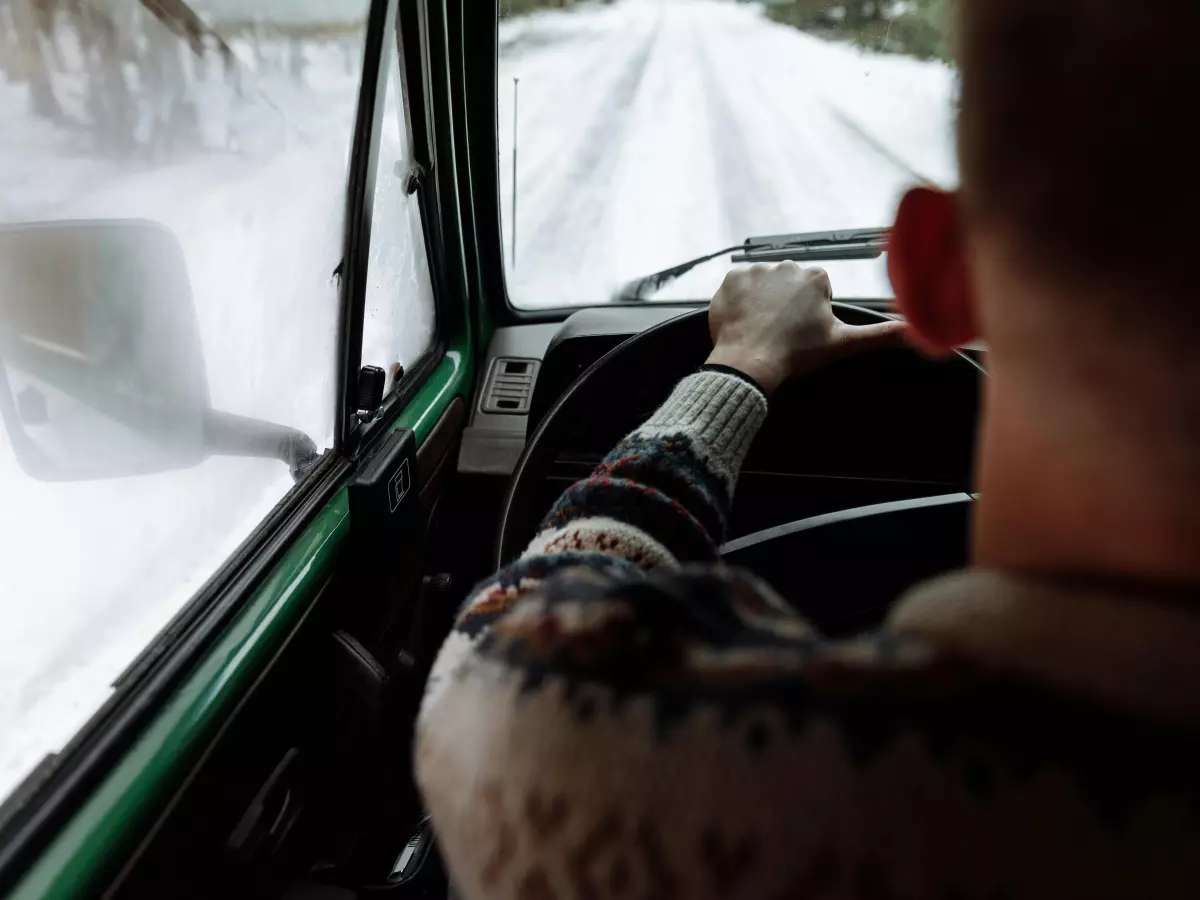 A person's arm and hand gripping a steering wheel, driving on a snowy road seen from inside the vehicle. 