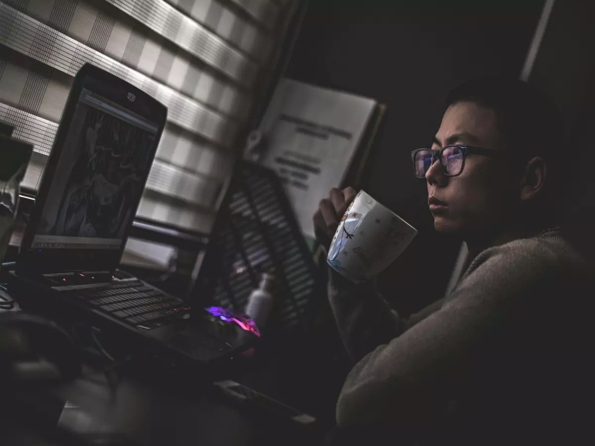 A person is sitting at a desk in a dimly lit room, working on a laptop. They are drinking coffee and looking at the screen.