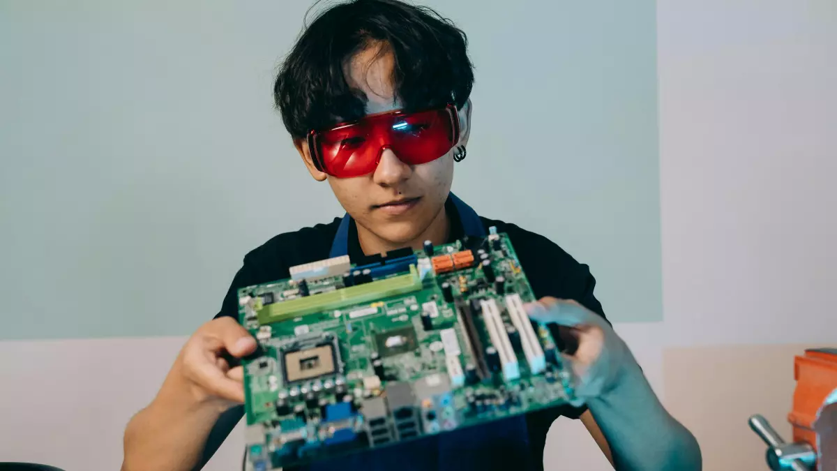 A person wearing red safety glasses holds a motherboard with a soldering iron on the table next to them.