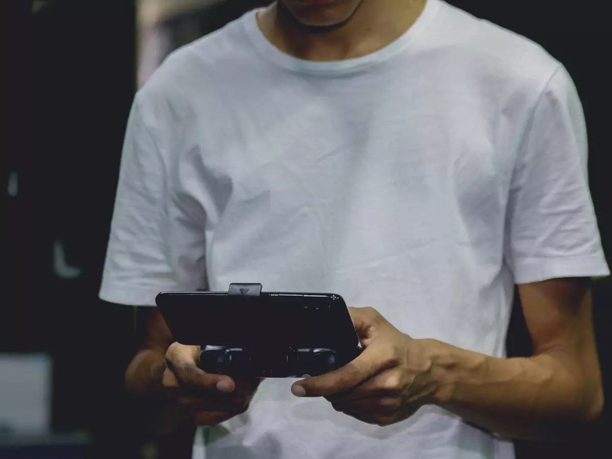 A man is holding a black smartphone in his hands. He is wearing a white t-shirt and is standing in a dark background.