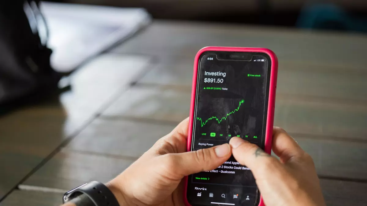 A person holding a smartphone with a bright screen showing a green line graph on a black background. The person