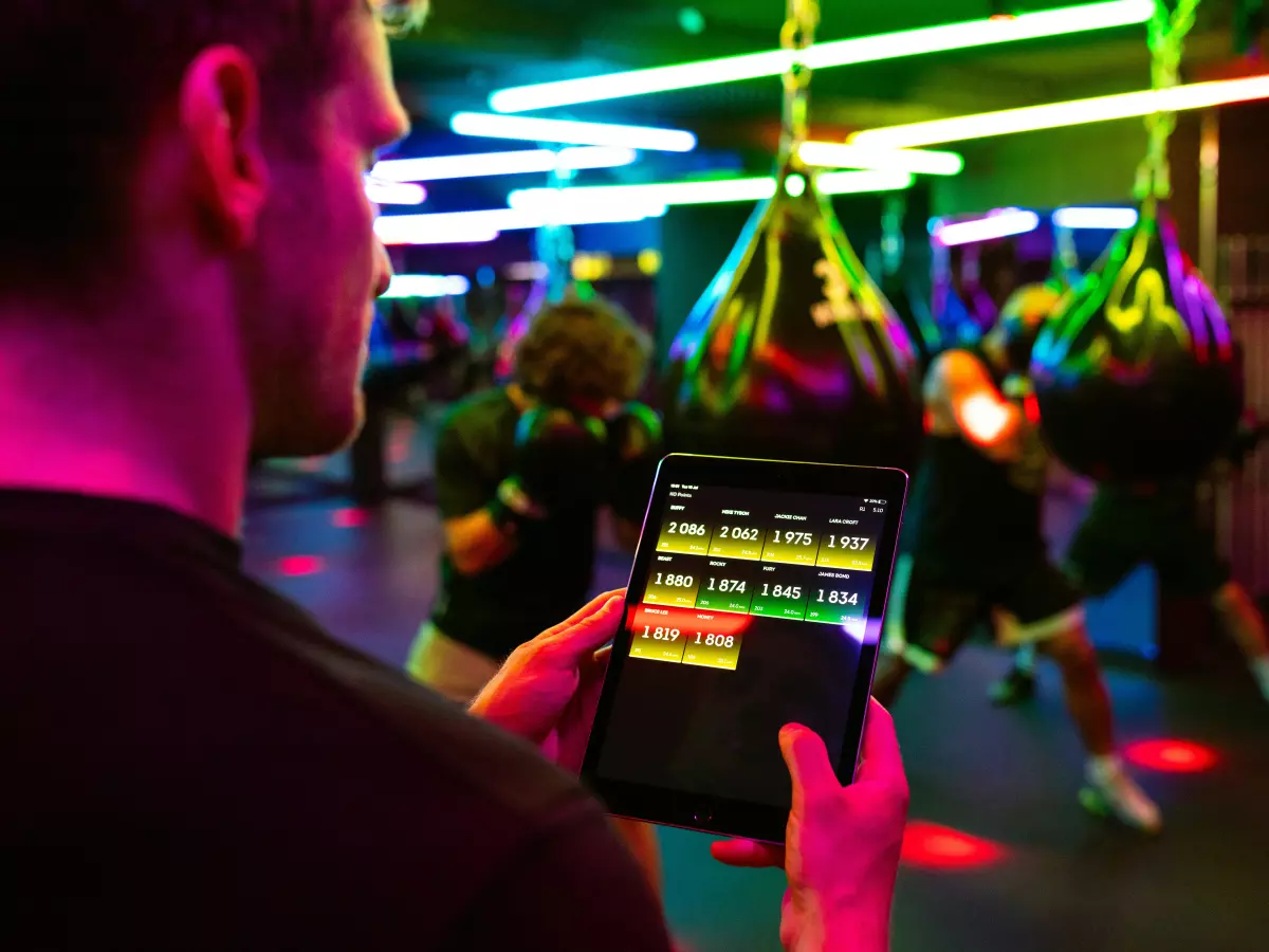 A man is using a tablet to track the performance of boxers in a boxing gym. There are multiple punching bags in the background. 