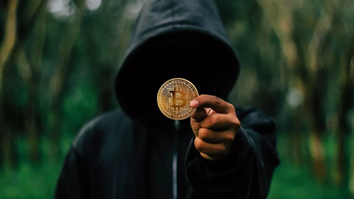 A person wearing a black hoodie is holding a bitcoin in their hand. They are standing in front of a blurred green background.