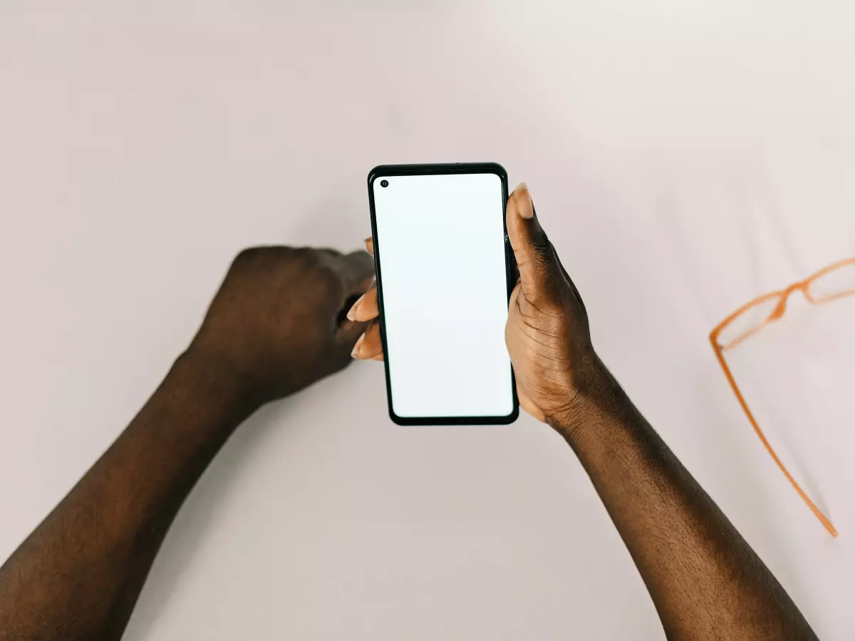 A person's hands are holding a smartphone with a white screen. The person's fingers are touching the screen. There are glasses lying on the table next to the phone.