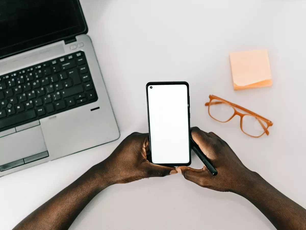 A person's hands holding a black smartphone with a white screen against a white background. A laptop, glasses, and a sticky note are also present on the table.