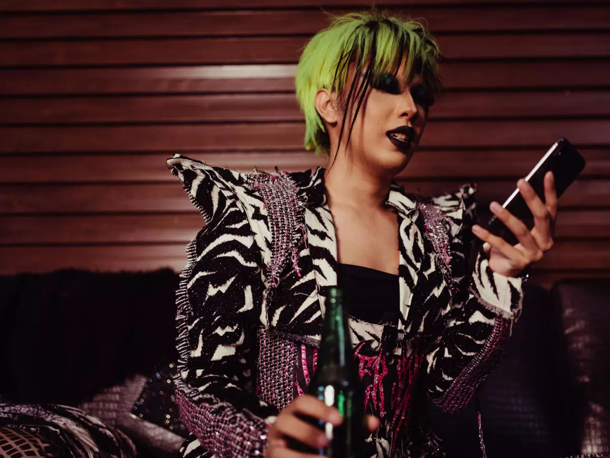 A person with green hair is sitting on a couch and using a smartphone. They are wearing a black and white jacket with spikes.