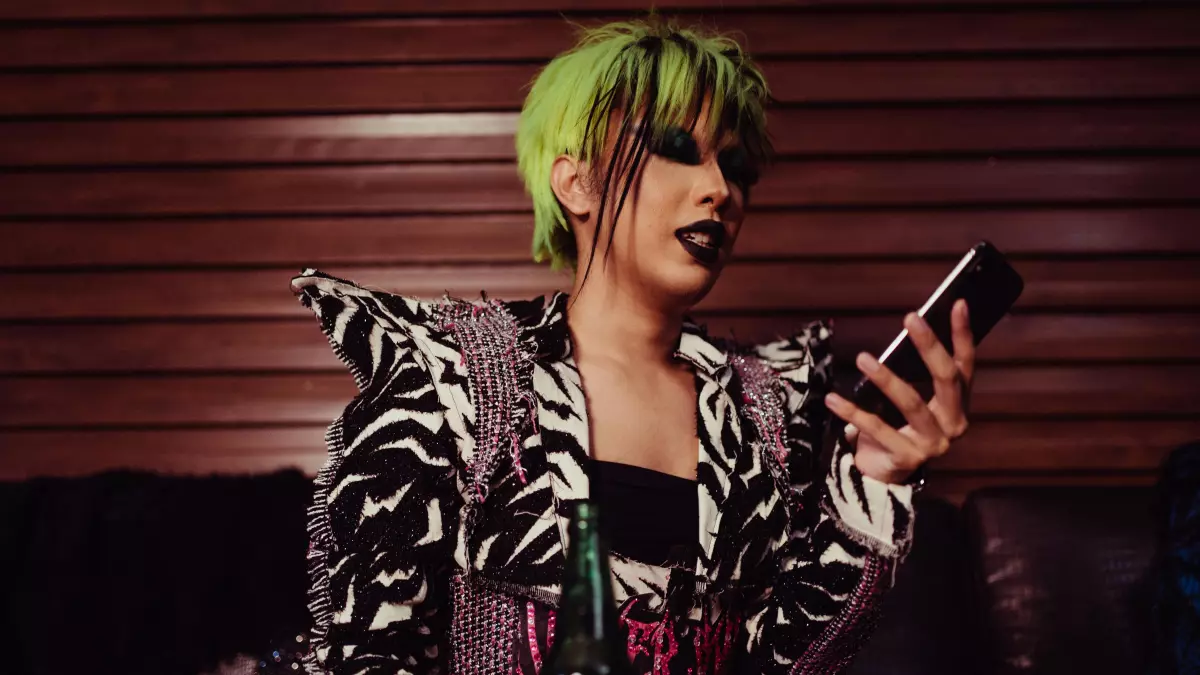 A person with green hair is sitting on a couch and using a smartphone. They are wearing a black and white jacket with spikes.